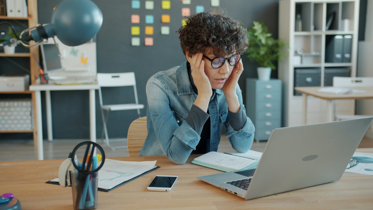 Office worker with glasses holds temples while looking at laptop.